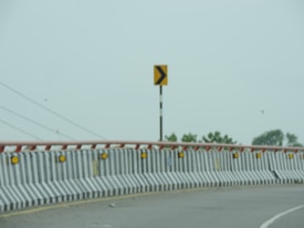A curved road with a concrete barrier featuring black and white stripes and yellow reflectors. A yellow and black directional sign is visible on a post. In the background, there are some trees and overhead wires against a cloudy sky.
