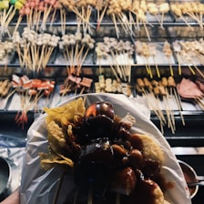 A variety of skewered foods is displayed on a rack, showcasing different meats and vegetables. In the foreground, a platter holds a serving of skewered items covered in a dark sauce with crispy chips on the side.