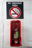 A sign indicating a no smoking area is positioned above a fire extinguisher enclosed in a red case, mounted on a white-painted wall.