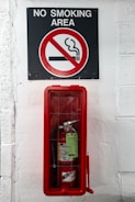 A sign indicating a no smoking area is positioned above a fire extinguisher enclosed in a red case, mounted on a white-painted wall.