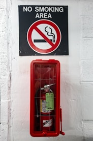 A sign indicating a no smoking area is positioned above a fire extinguisher enclosed in a red case, mounted on a white-painted wall.