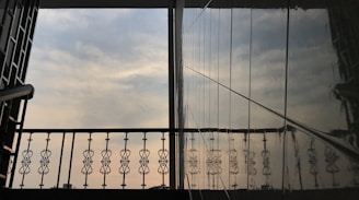 An architectural view featuring a decorative metal railing against the backdrop of a cloudy sky. The reflection of the railing is visible on a glossy wall surface, creating a symmetrical and artistic visual. The scene suggests it might be from a balcony or a similar elevated structure.