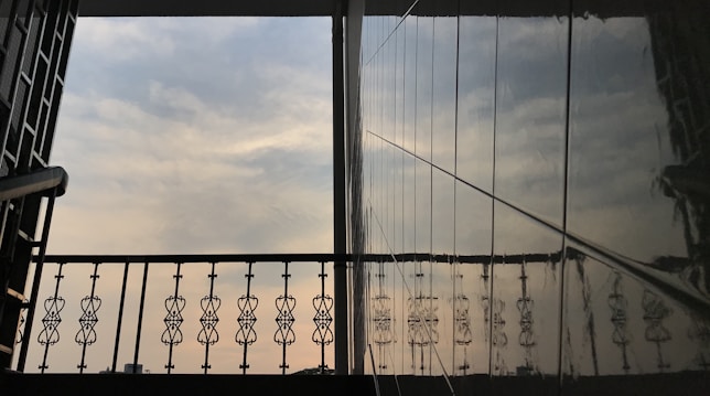 An architectural view featuring a decorative metal railing against the backdrop of a cloudy sky. The reflection of the railing is visible on a glossy wall surface, creating a symmetrical and artistic visual. The scene suggests it might be from a balcony or a similar elevated structure.