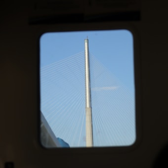 A bridge with a tall, sleek metallic tower is seen through a window, with multiple cables extending from the tower against a clear blue sky.