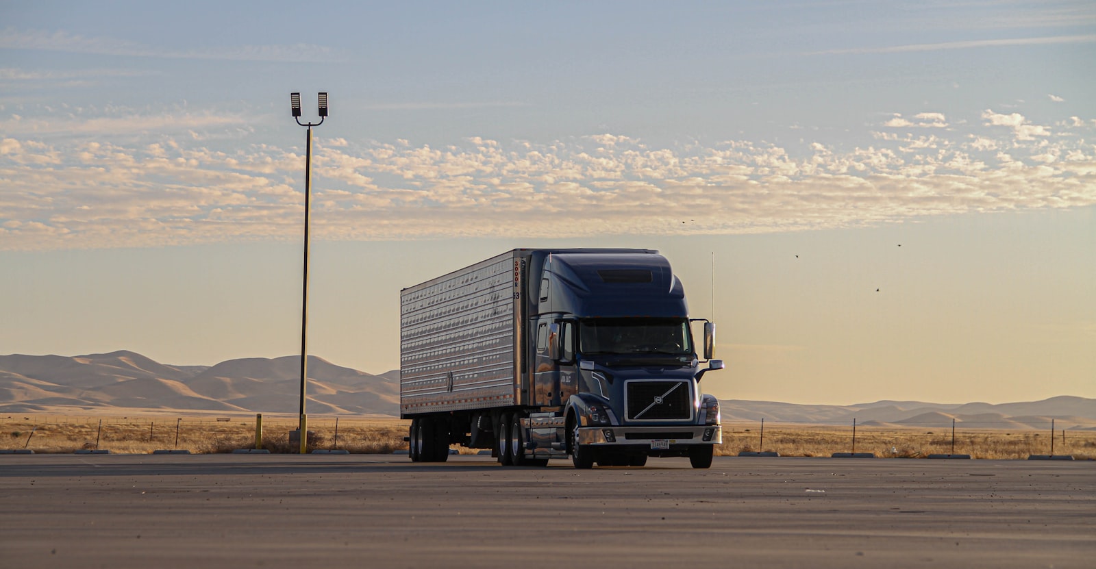 Dark blue semi-trailer truck parked on asphalt lot with rolling hills and warm sky in background