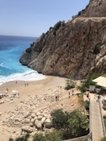 A coastal scene featuring a sandy beach bordered by rugged cliffs with a few people walking around. The turquoise waves gently lap against the shore, and there are several lounge chairs and umbrellas scattered across the beach. A wooden staircase leads down to the sand from a nearby path surrounded by lush green plants.