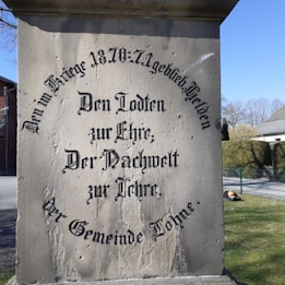 Old monument with engraved text in German, located outdoors amidst buildings and greenery, including manicured bushes, a tree, and a street.