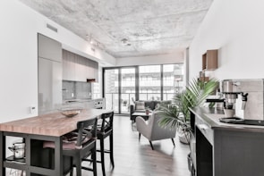 A modern open-concept living space features a sleek kitchen with stainless steel appliances and a large wooden island equipped with bar stools. A cozy seating area with a sofa and armchair overlooks large floor-to-ceiling windows, allowing natural light to flood the space. Indoor plants add a touch of greenery, and contemporary decor pieces accentuate the minimalist aesthetic.