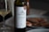 A bottle of Robola of Kefalonia white wine is positioned prominently on a table. The label on the bottle is clearly visible, featuring a detailed illustration of a temple-like structure. Beside the bottle, there is a glass containing white wine. In the background, a plate with cooked prawns is partially visible, suggesting a meal setting.