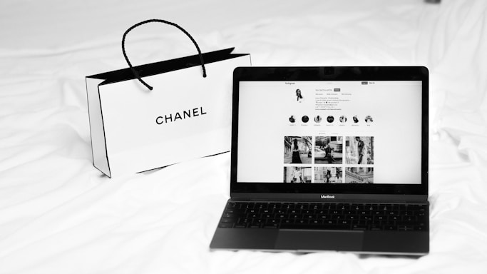 A black laptop displays a social media profile on its screen, placed on a white bed next to a Chanel shopping bag