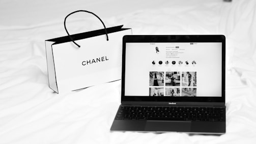 A black laptop displays a social media profile on its screen, placed on a white bed next to a Chanel shopping bag