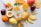 A colorful assortment of fruits arranged on a light surface, including slices of orange, apple, passion fruit, and mango. A bottle of mango juice with the label 'Natural One' sits prominently in the center alongside a glass of orange juice.