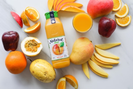 A colorful assortment of fruits arranged on a light surface, including slices of orange, apple, passion fruit, and mango. A bottle of mango juice with the label 'Natural One' sits prominently in the center alongside a glass of orange juice.