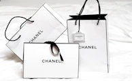 Three white shopping bags with black handles and bold black 'Chanel' branding displayed prominently. The bags are set against a light background, creating a minimalist and luxurious aesthetic. One of the bags has a small tag with additional branding.