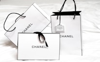 Three white shopping bags with black handles and bold black 'Chanel' branding displayed prominently. The bags are set against a light background, creating a minimalist and luxurious aesthetic. One of the bags has a small tag with additional branding.