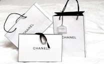 Three white shopping bags with black handles and bold black 'Chanel' branding displayed prominently. The bags are set against a light background, creating a minimalist and luxurious aesthetic. One of the bags has a small tag with additional branding.