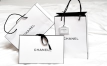 Three white shopping bags with black handles and bold black 'Chanel' branding displayed prominently. The bags are set against a light background, creating a minimalist and luxurious aesthetic. One of the bags has a small tag with additional branding.