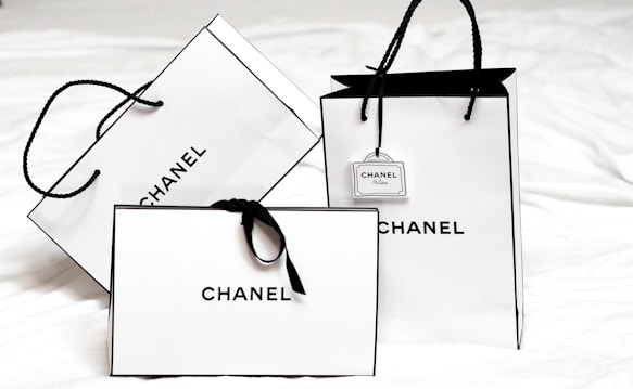 Three white shopping bags with black handles and bold black 'Chanel' branding displayed prominently. The bags are set against a light background, creating a minimalist and luxurious aesthetic. One of the bags has a small tag with additional branding.