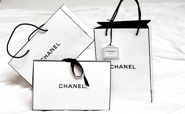 Three white shopping bags with black handles and bold black 'Chanel' branding displayed prominently. The bags are set against a light background, creating a minimalist and luxurious aesthetic. One of the bags has a small tag with additional branding.