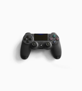 A black video game controller with various buttons and dual analog sticks, placed against a white background. It includes iconic PlayStation symbols such as triangles, circles, crosses, and squares on its buttons.