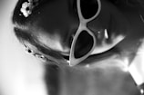 A woman is wearing large, white-framed cat-eye sunglasses. Her hair is adorned with small white flowers. The image is in black and white, with a focus on her face and accessories.