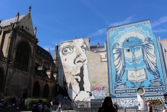 A large mural featuring a monochrome portrait of a face with a finger on its lips conveys a message of silence or secrecy. Next to it, another mural in shades of blue shows a stylized image with two figures holding a blooming flower over an open book, accompanied by the words 'THE FUTURE IS UNWRITTEN'. Both murals are painted on the side of adjacent buildings with gothic architectural elements visible on the left.