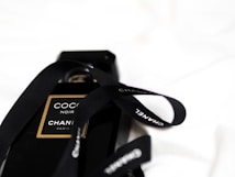 A black bottle of Coco Noir perfume by Chanel is adorned with a black ribbon featuring the brand name. The bottle and ribbon are set against a soft, white background, creating a contrast that highlights the elegant design.