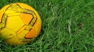 A vibrant yellow soccer ball with black patterns is resting on lush green grass. The ball is slightly worn, indicating it has been used for playing outdoors.