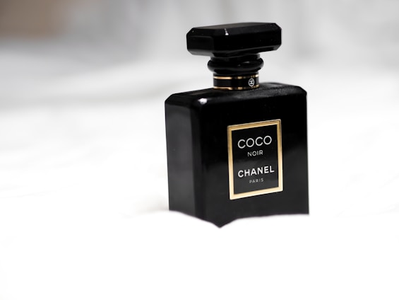 A sleek black perfume bottle with a geometric design, displaying a gold-edged label that reads 'Coco Noir Chanel Paris.' It is set against a soft, blurred white background, emphasizing its luxurious and elegant appearance.