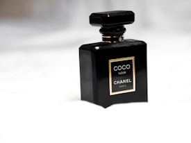 A sleek black perfume bottle with a geometric design, displaying a gold-edged label that reads 'Coco Noir Chanel Paris.' It is set against a soft, blurred white background, emphasizing its luxurious and elegant appearance.
