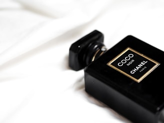 A black perfume bottle labeled 'COCO NOIR CHANEL PARIS' is placed on a smooth, white fabric background. The design is sleek and elegant with gold accents around the label.