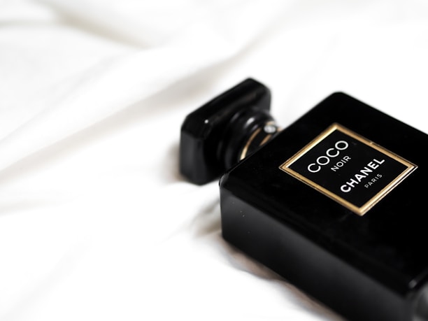 A black perfume bottle labeled 'COCO NOIR CHANEL PARIS' is placed on a smooth, white fabric background. The design is sleek and elegant with gold accents around the label.