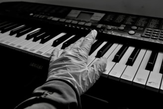 A gloved hand is placed on the keys of a keyboard, suggesting careful interaction or play. The keyboard appears to be a digital musical instrument, with visible control buttons and a display screen. The monochrome setting creates a stark contrast between the black and white keys.