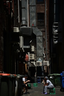 An urban alleyway scene with two people sitting on crates conversing. The alley is lined with air conditioning units and pipes, and there is graffiti on the walls. Trash bins are visible along the sides, and the lighting creates strong contrasts with patches of shadow and sunlight.