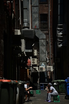 An urban alleyway scene with two people sitting on crates conversing. The alley is lined with air conditioning units and pipes, and there is graffiti on the walls. Trash bins are visible along the sides, and the lighting creates strong contrasts with patches of shadow and sunlight.