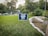 A yard sign with the message 'I'M SO GONNA VOTE' and the date 'November 3, 2020' stands on a grassy lawn. In the background, there are trees and a suburban neighborhood with parked cars and houses. The sign is supported by two slim metal stakes. A stone staircase and pathway are visible to the right.