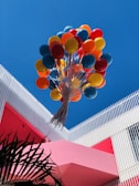 A vibrant bunch of colorful balloons, including red, orange, yellow, and blue, floats upward against a clear blue sky. The balloons are anchored to a structure featuring modern architectural elements, with white vertical slats and a bold pink wall. Palm leaves are visible in the lower left corner.