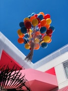 A vibrant bunch of colorful balloons, including red, orange, yellow, and blue, floats upward against a clear blue sky. The balloons are anchored to a structure featuring modern architectural elements, with white vertical slats and a bold pink wall. Palm leaves are visible in the lower left corner.