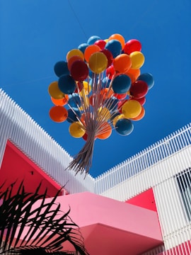 A vibrant bunch of colorful balloons, including red, orange, yellow, and blue, floats upward against a clear blue sky. The balloons are anchored to a structure featuring modern architectural elements, with white vertical slats and a bold pink wall. Palm leaves are visible in the lower left corner.