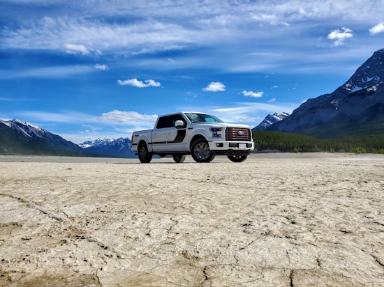 A white pickup truck is parked on a cracked, dry landscape with rugged mountains and a clear blue sky in the background. The scene conveys a sense of vastness and openness, with the natural environment highlighting the vehicle.