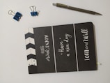 A black notebook with motivational phrases written in white font on the cover is placed on a light surface. There are two small blue binder clips and one larger blue binder clip nearby, along with a silver pen.