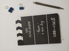 A black notebook with motivational phrases written in white font on the cover is placed on a light surface. There are two small blue binder clips and one larger blue binder clip nearby, along with a silver pen.