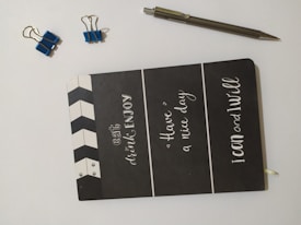 A black notebook with motivational phrases written in white font on the cover is placed on a light surface. There are two small blue binder clips and one larger blue binder clip nearby, along with a silver pen.