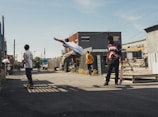 Several young people are skateboarding in an urban alleyway. One person is performing an airborne trick while others watch or prepare to skate. The scene has a casual and relaxed atmosphere with industrial buildings and clear skies in the background.
