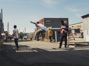 Several young people are skateboarding in an urban alleyway. One person is performing an airborne trick while others watch or prepare to skate. The scene has a casual and relaxed atmosphere with industrial buildings and clear skies in the background.