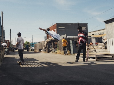 Several young people are skateboarding in an urban alleyway. One person is performing an airborne trick while others watch or prepare to skate. The scene has a casual and relaxed atmosphere with industrial buildings and clear skies in the background.
