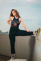 A woman with long, wavy hair is posing confidently on an urban rooftop. She is wearing a sleeveless top with the phrase 'Live Life Loud' and black sweatpants. One of her legs is raised and resting on a ledge, showing off her sneakers. The background features a cityscape under a soft, cloudy sky.