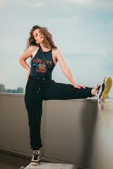 A woman with long, wavy hair is posing confidently on an urban rooftop. She is wearing a sleeveless top with the phrase 'Live Life Loud' and black sweatpants. One of her legs is raised and resting on a ledge, showing off her sneakers. The background features a cityscape under a soft, cloudy sky.