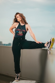 A woman with long, wavy hair is posing confidently on an urban rooftop. She is wearing a sleeveless top with the phrase 'Live Life Loud' and black sweatpants. One of her legs is raised and resting on a ledge, showing off her sneakers. The background features a cityscape under a soft, cloudy sky.