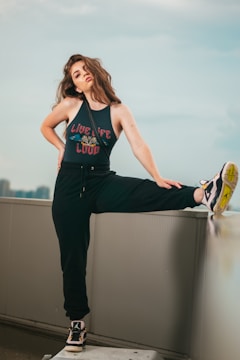 A woman with long, wavy hair is posing confidently on an urban rooftop. She is wearing a sleeveless top with the phrase 'Live Life Loud' and black sweatpants. One of her legs is raised and resting on a ledge, showing off her sneakers. The background features a cityscape under a soft, cloudy sky.